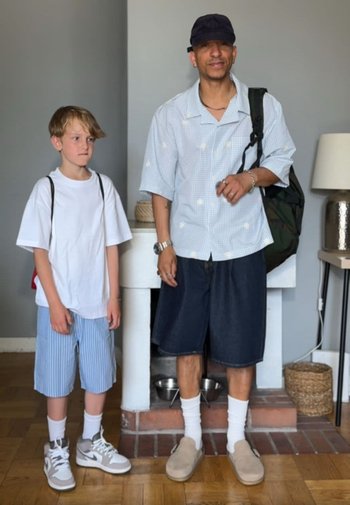 Un jeune garçon et un homme adulte se tiennent à l’intérieur, tous deux portent des shorts décontractés, des chaussettes blanches et des sacs à dos, devant un mur gris clair et une cheminée.