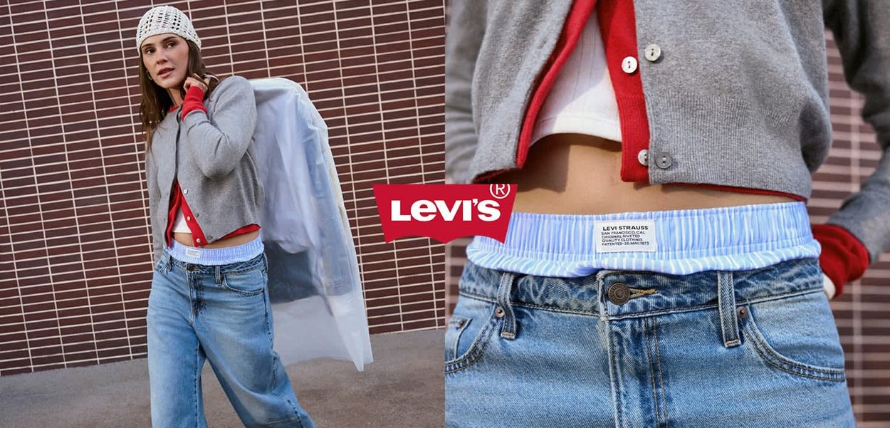 Frau trägt geschichtete graue und rote Strickjacken, blaue Jeans mit gestreiftem Bund, hält eine weiße Kleidertasche vor einer gefliesten Wand.