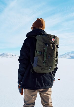 Grüner Canvas-Rucksack mit Roll-Top-Design, verstellbaren Trägern, grauen Akzenten und einem vorderen Bungee-Kordel. Tragen von einer Person in Winterkleidung.