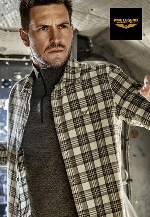 Geruite overhemd in beige en bruin, gedragen over een grijze top met hoge hals. Voorzien van knoopsluitingen en subtiel logodetail op het overhemd.