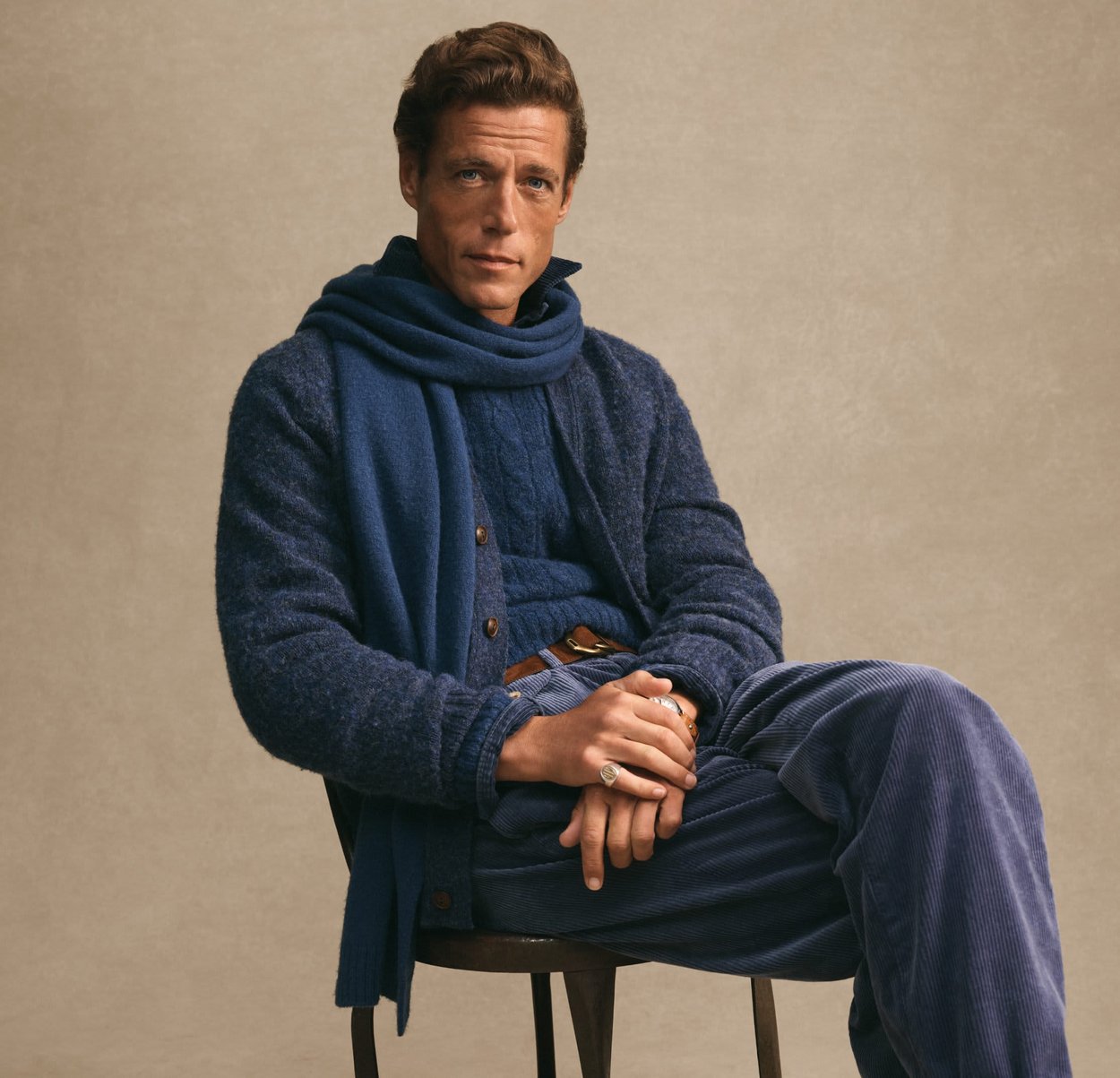 Uomo seduto su una sedia, indossando un maglione a strati blu, una sciarpa e pantaloni in velluto a coste, su uno sfondo beige uniforme.