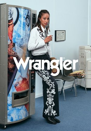 Zwarte denim broeken met witte bloemenborduursels langs de zijkanten. Het model draagt een witte overhemden met zwarte accenten en staat naast een automaat.