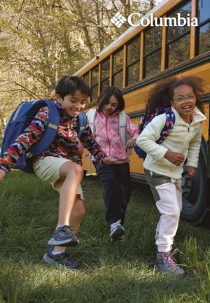 Three children with backpacks cheerfully step off a yellow school bus onto grass, wearing colorful outdoor jackets in a wooded area.