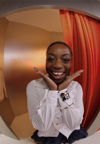 Femme souriante encadrant son visage avec ses mains, portant une chemise blanche et des bracelets argentés, debout devant un rideau orange et un mur beige.