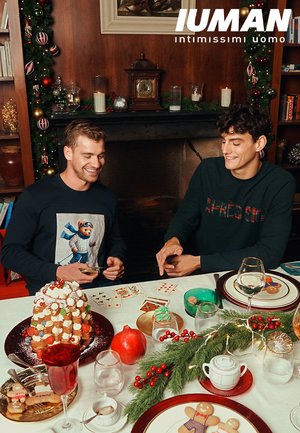 Deux hommes assis à une table festive. L'un porte un pull bleu marine avec un motif d'ours ; l'autre porte un pull vert foncé. Un dessert coloré et des décorations de fête sont présents.