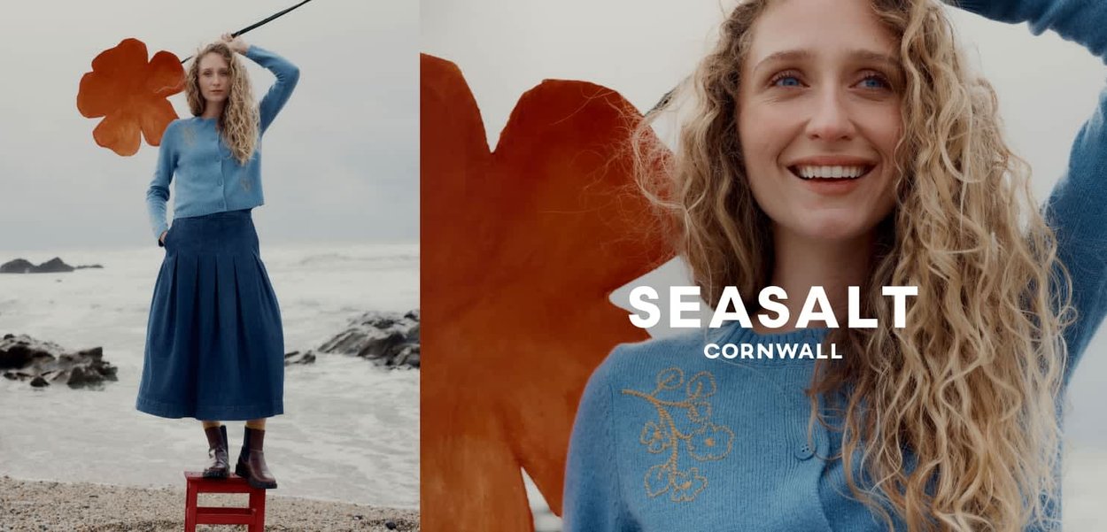 Frau mit lockigem Haar, die einen blauen Pullover und einen Rock trägt, steht auf einem roten Hocker an einem felsigen Strand und hält ein orangefarbenes Blatt hinter sich.