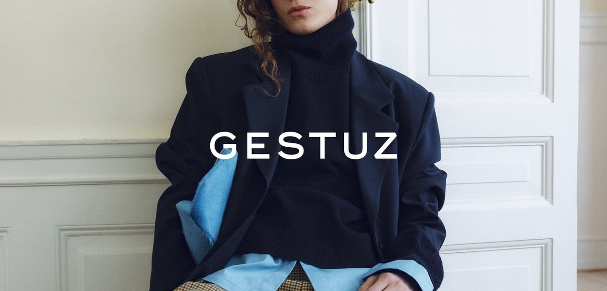 Vrouw draagt een gelaagde oversized zwarte blazer, blauw overhemd, zwarte coltrui en gebloemde broek, zittend tegen een witte wand met panelen en de tekst "GESTUZ".