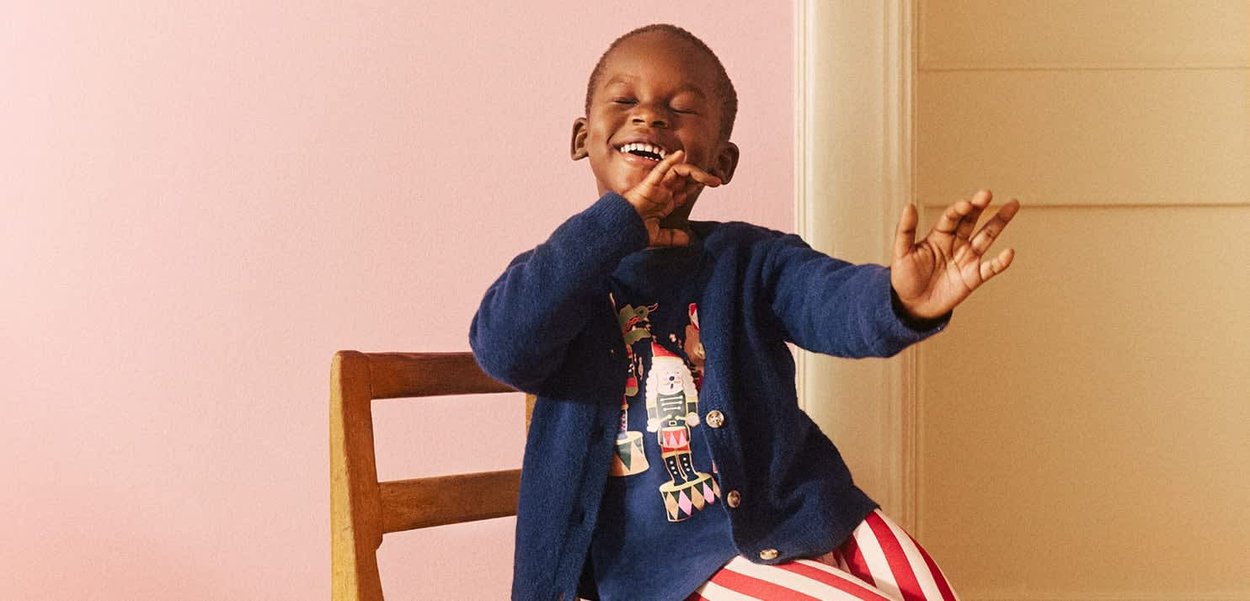 Dziecko w niebieskim kardiganie na wzorzystej koszuli z motywem szachowego króla i w paskowanych spodniach, siedzące na drewnianym krześle przed różowym tłem.