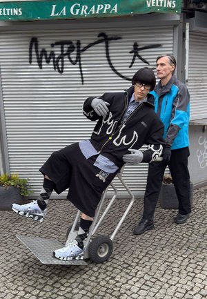 Metallkarre mit zwei Rädern in aufrechter Haltung. Eine Person in schwarzer Jacke und Shorts, mit Handschuhen, balanciert lässig darauf. Grauer Asphalt.