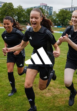 Three female soccer players in black uniforms celebrating and running on a grass field with buildings in the background.