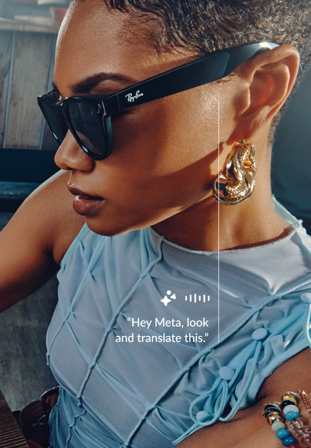 Lunettes de soleil noires avec des montures épaisses, un haut texturé bleu clair et une boucle d'oreille abstraite dorée. Un bracelet au poignet comporte des perles colorées.