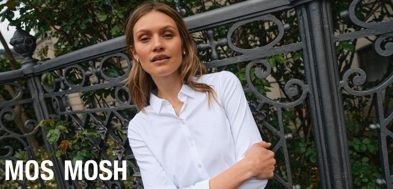 Woman in a white button-up shirt stands with arms crossed in front of decorative black iron fence and green foliage.