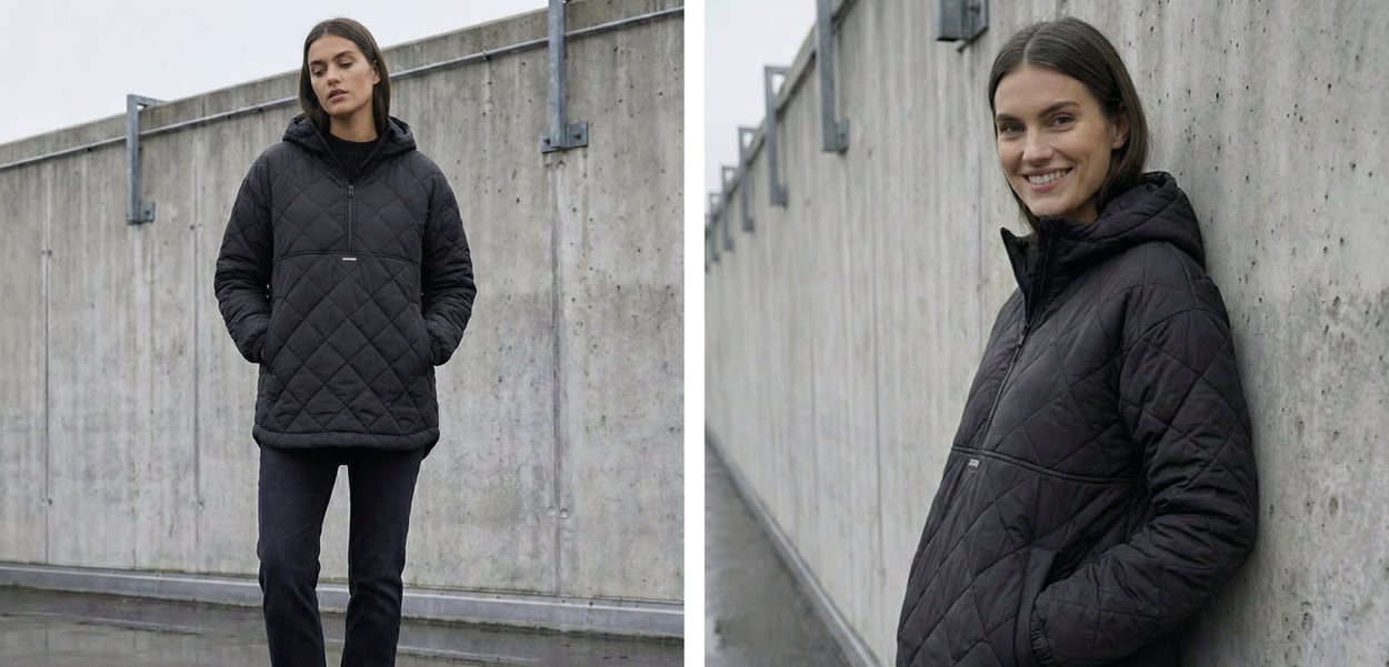 Woman in black quilted jacket and dark pants stands and leans against a concrete wall outdoors with a neutral and smiling expression.