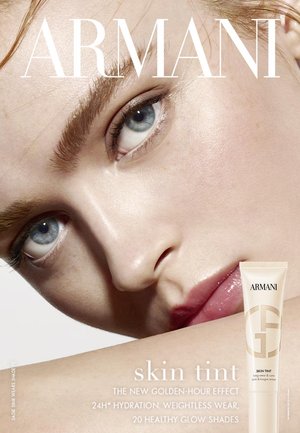 Nahaufnahme eines Gesichts mit glatter Haut, blauen Augen und natürlicher Schminke. Eine beige Produkttube mit der Aufschrift "Armani Skin Tint" ist teilweise sichtbar.