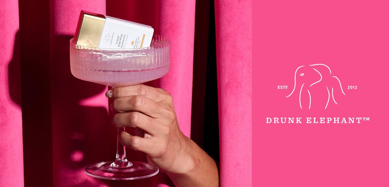 Klares Glascoupe hält ein Hautpflegeprodukt mit einem goldenen Verschluss, vor einem lebhaften pinkfarbenen Hintergrund mit strukturierten Vorhängen platziert.