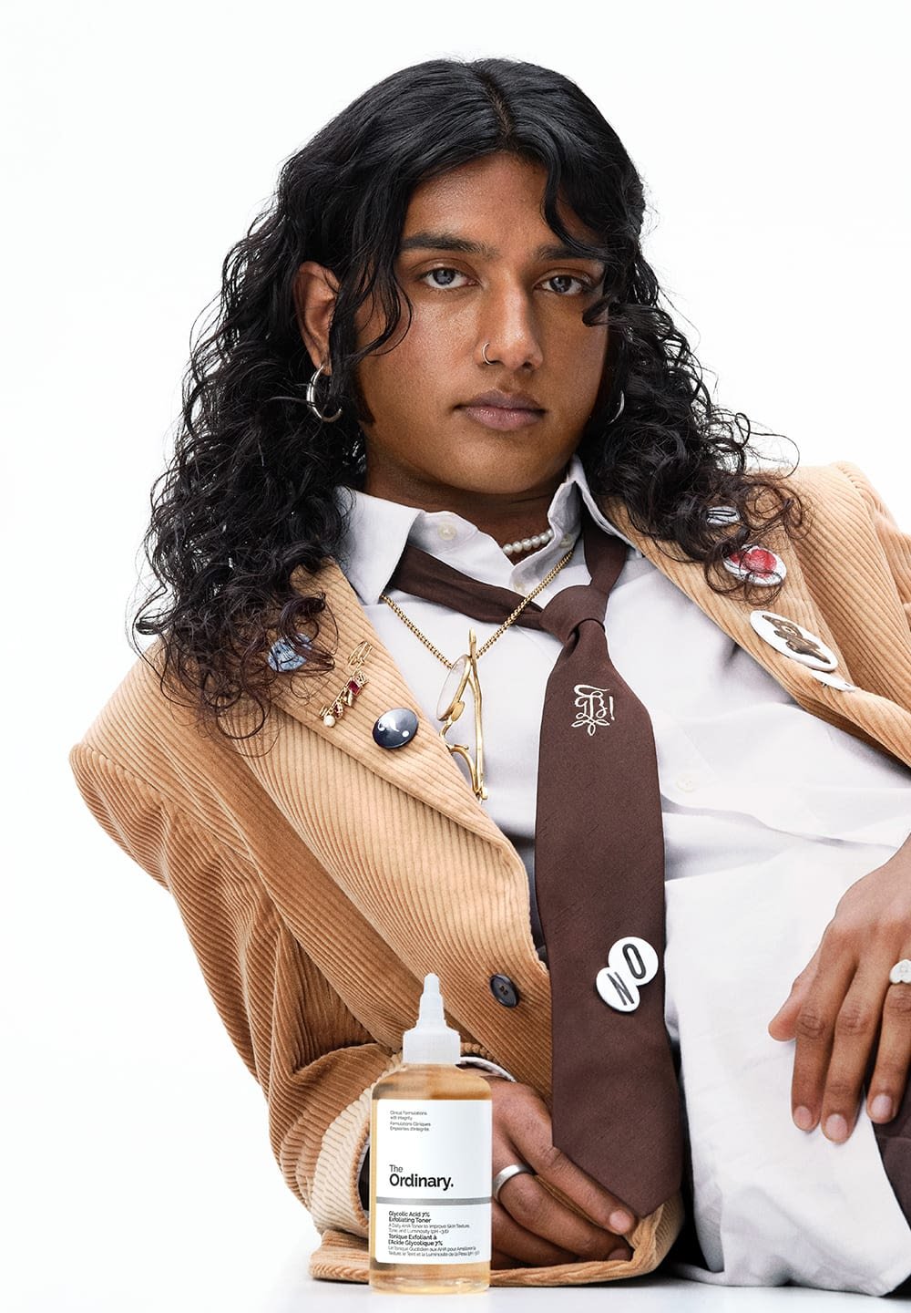A model with curly black hair wears a tan corduroy jacket and a brown tie, accessorized with pins and a pearl necklace, holding a skincare product.