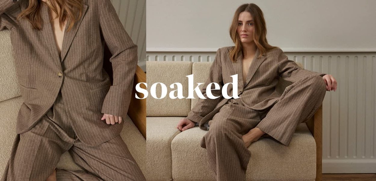 Woman in brown pinstripe suit sitting barefoot on beige sofa against white wall with radiator, word "soaked" overlaid.