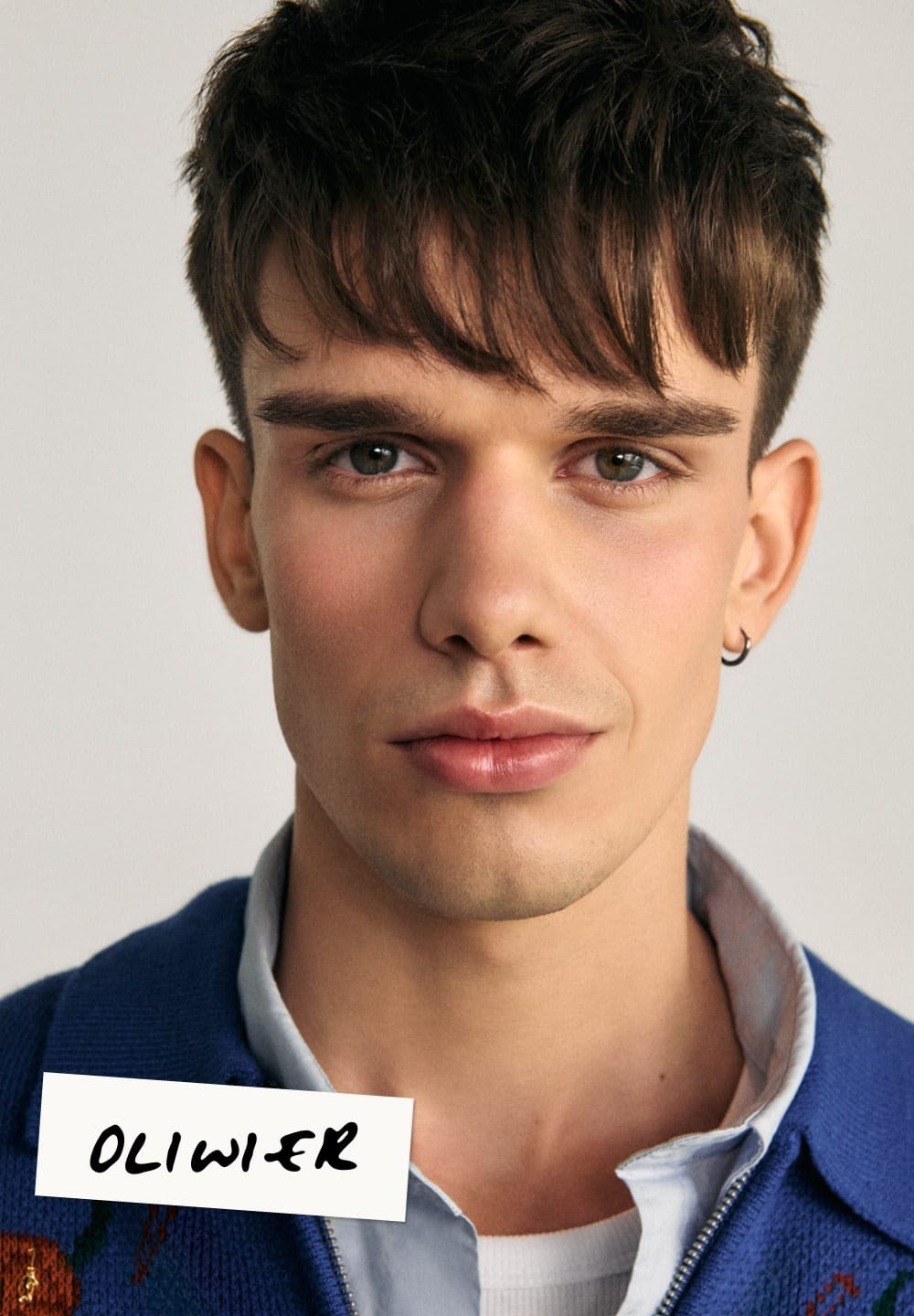 Zbliżenie młodego mężczyzny z krótkimi ciemnymi włosami, noszącego niebieski kardigan na jasnej koszuli, z przekutą uchem i gładką skórą.