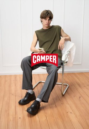 Green sleeveless shirt, gray pinstripe trousers, black shoes with thick soles, and light gray socks. Model seated in a metal chair.