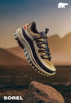 Braune und schwarze Wanderschuhe mit aggressivem Profil, die über einem Felsen in einer bergigen Umgebung bei Sonnenuntergang schwebt.