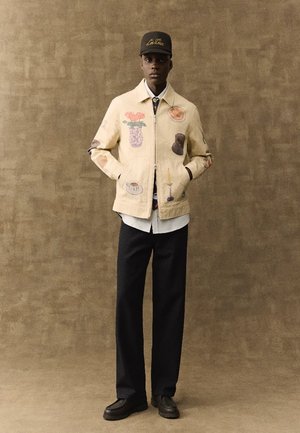 Mand iført beige jakke med farverige lapper, hvid skjorte, sorte bukser, sorte loafers og sort kasket mod ensfarvet brun baggrund.