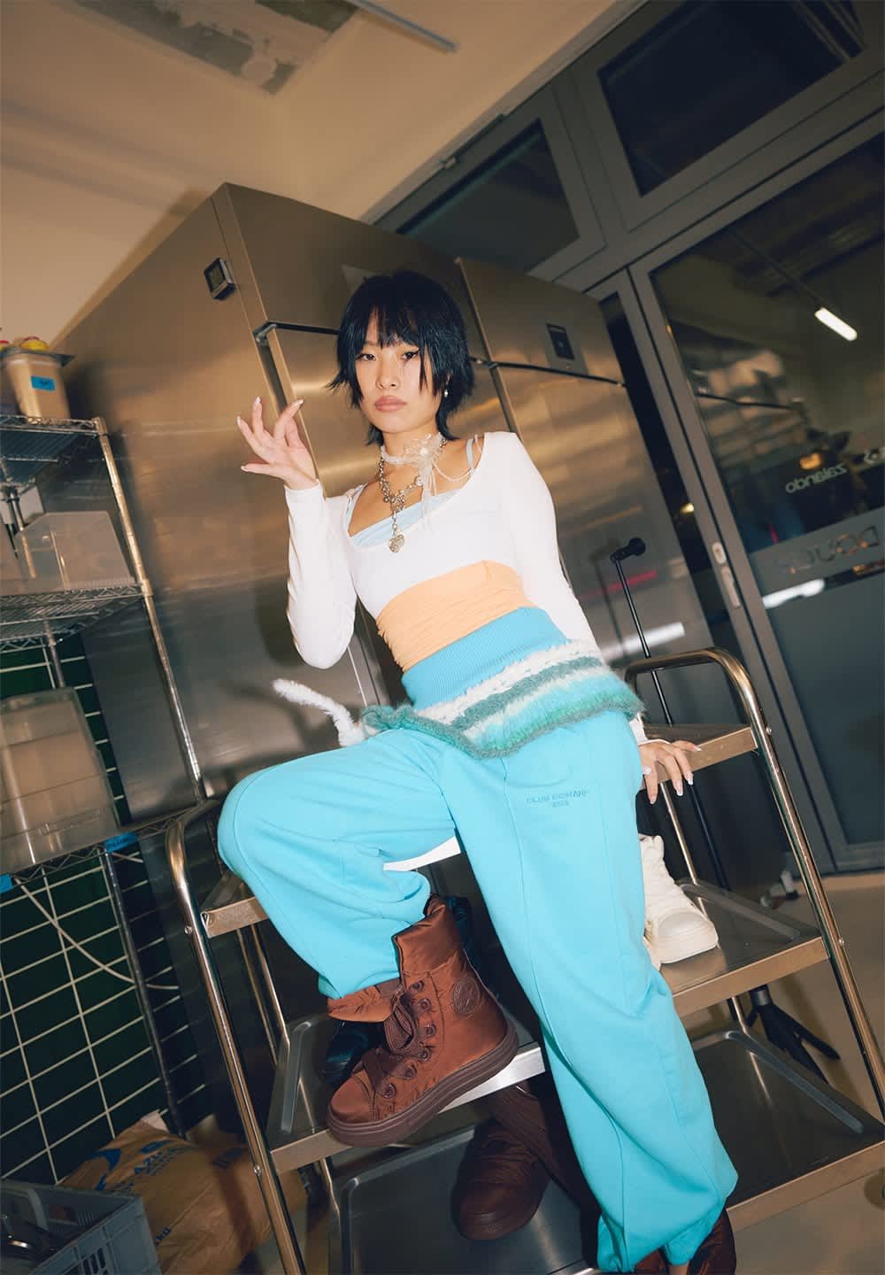 La modelo lleva una camiseta blanca de manga larga sobre un top color durazno, combinada con pantalones deportivos turquesa y botas marrones con cordones, en un ambiente de cocina.