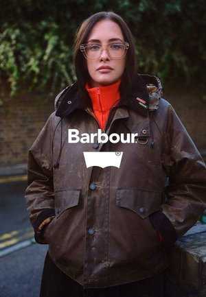 Brown waxed jacket with a black collar, button closures, and zipped pockets. Layered with a bright red inner lining for contrast.