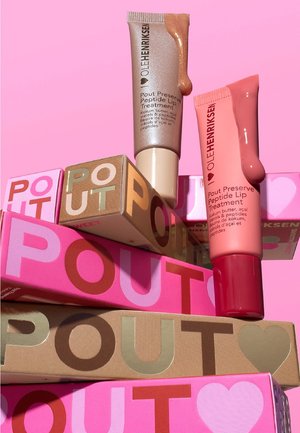 Due tubetti di Ole Henriksen Pout Preserve Peptide Lip Treatment con campioni di colore, circondati da scatole colorate impilate del prodotto POUT su uno sfondo rosa.