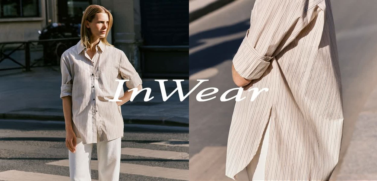 Kvinde iført en beige stribet oversized skjorte og hvide bukser, stående udendørs på en bygade i sollys.