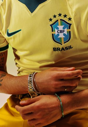 Persona con una camiseta de fútbol amarilla de Brasil, joyas en ambas muñecas, brazos cruzados sobre el cuerpo y un tatuaje visible en el brazo izquierdo.