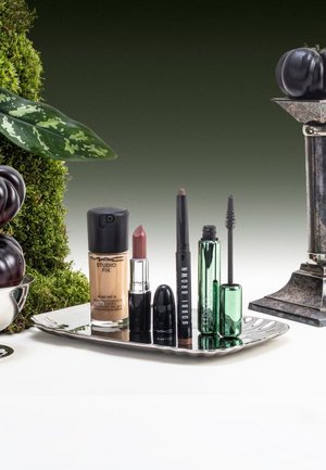 Collezione di prodotti cosmetici su un vassoio d'argento: fondotinta MAC, rossetto, matita Bobbi Brown, mascara verde, con uno sfondo decorativo.