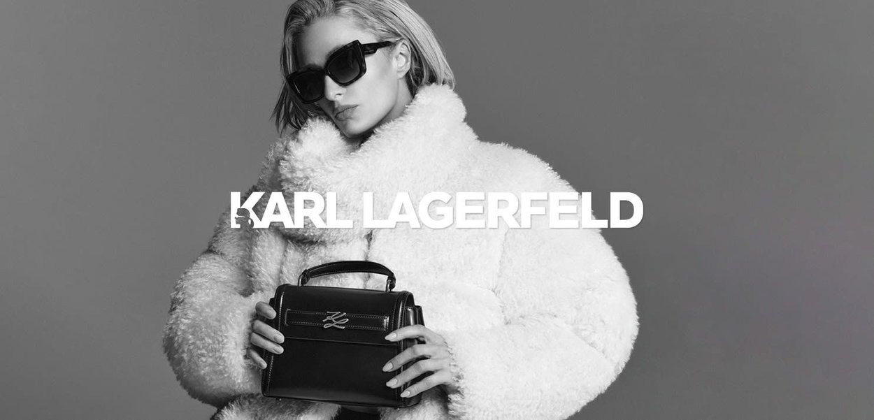 Black leather handbag with a structured design and silver hardware, held in hands. Model wears a fluffy white coat and dark sunglasses.