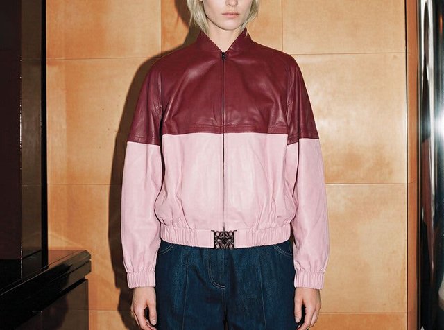 Personne portant une veste en cuir bicolore en bordeaux et rose clair, associée à un pantalon en jean bleu foncé, debout contre un mur carrelé.