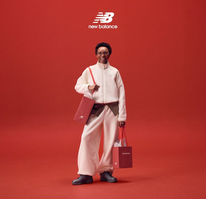 Giacca con cerniera in pile bianco e pantaloni abbinati, scarpe grigie, che tengono borse della spesa rosse, su uno sfondo rosso uniforme.