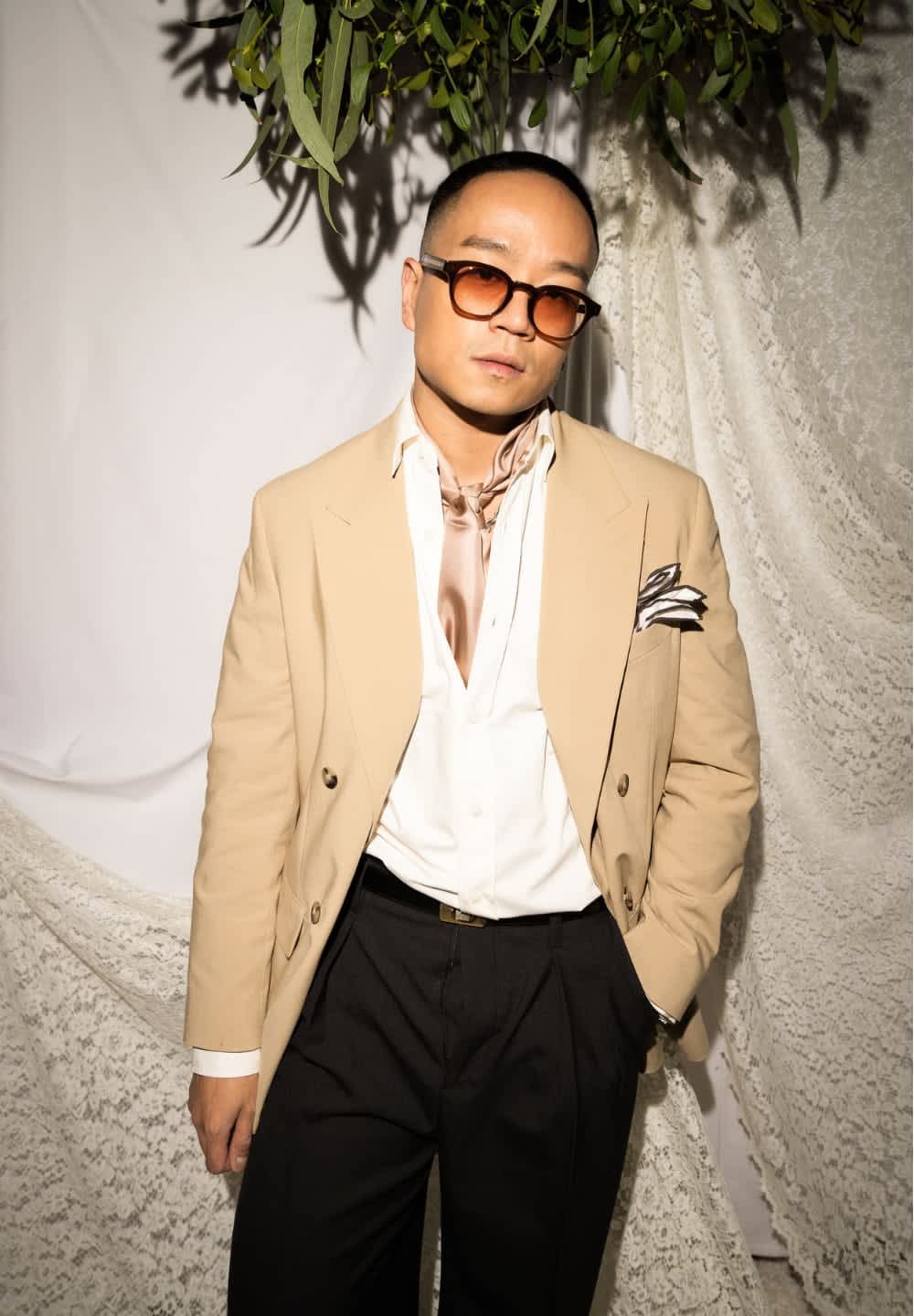 Beige dubbelknäppt blazer över en vit skjorta, krämfärgad halsduk, svarta byxor och svarta glasögon, mot en strukturerad vit bakgrund.