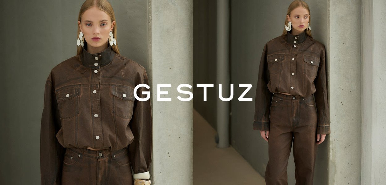 Ung kvinde iført mørkebrun denimjakke med knapper og matchende højtaljede jeans, stående ved lysegrå betonpiller.