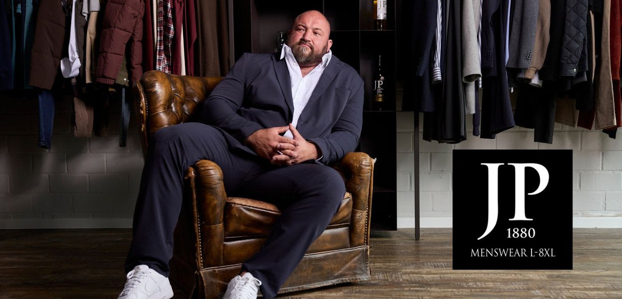 Marineblauer Anzug mit weißem Hemd, sitzend auf einem braunen Ledersessel. Im Hintergrund hängt ein Ständer mit verschiedenen Kleidungsstücken. Schwarz-weißes Logo sichtbar.