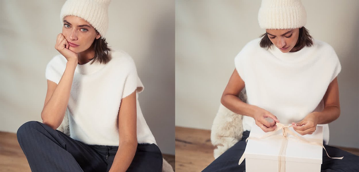 Chapeau blanc tricoté et pull blanc duveteux à manches courtes avec encolure arrondie. Assis et en train de dénouer un ruban sur une boîte cadeau blanche.