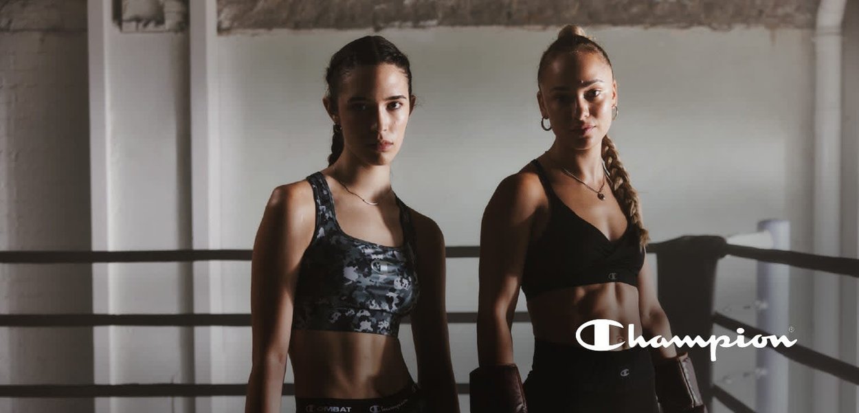 Dos mujeres con camisetas deportivas están en un ring de boxeo. Una lleva una camiseta negra con camuflaje, la otra una camiseta negra lisa. Ambas tienen guantes de boxeo.