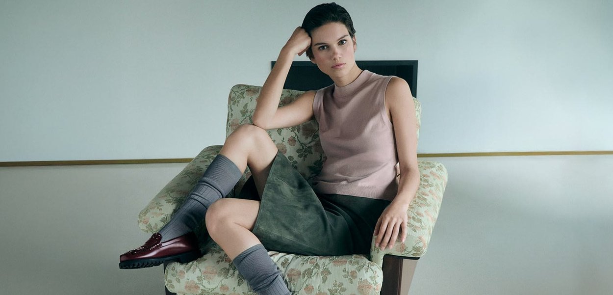 Knit sleeveless top in light pink, olive green suede skirt, gray knee-high socks, and burgundy shoes, seated on a floral-patterned chair.