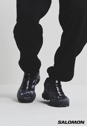 Person i sorte Salomon sneakers og sorte bukser, der står på et lys grå gulv, vist fra midt på læggen og ned.