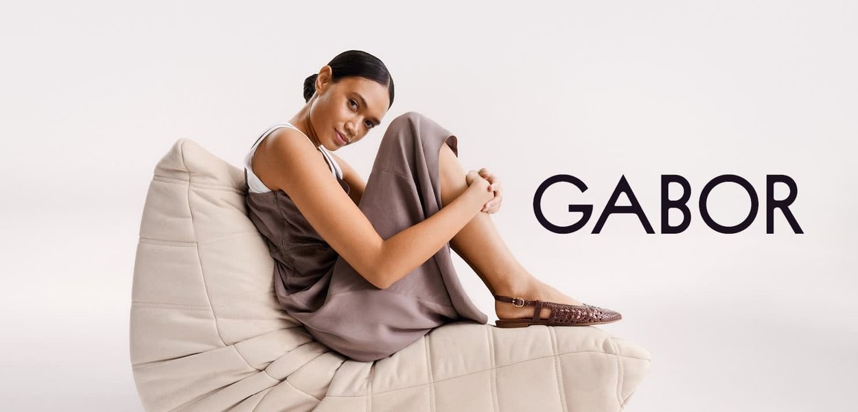 Vrouw in een bruine jurk en gevlochten platte schoenen, zittend op een zachte, beige gevoerde stoel tegen een effen witte achtergrond met de tekst "GABOR".