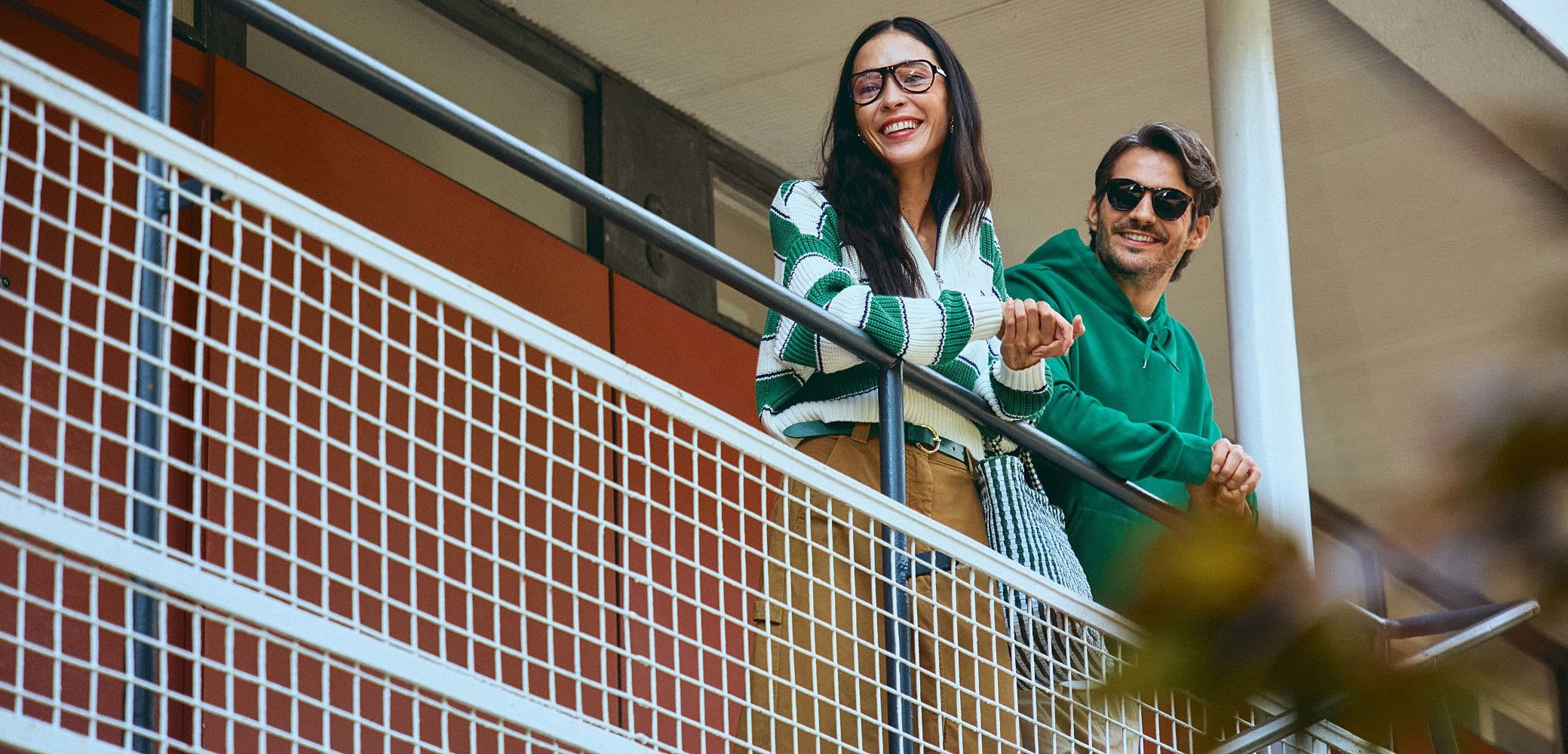 Due persone sono in piedi su un balcone. Una indossa un maglione a righe verde e bianco con occhiali, mentre l'altra indossa un felpa verde e occhiali da sole.