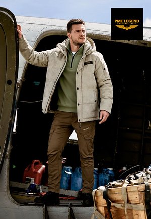 Lichtgrijze geïsoleerde jas met een capuchon over een groene lange mouwen top, gecombineerd met beige cargobroek en zwarte schoenen. Staande in een vliegdeuren.