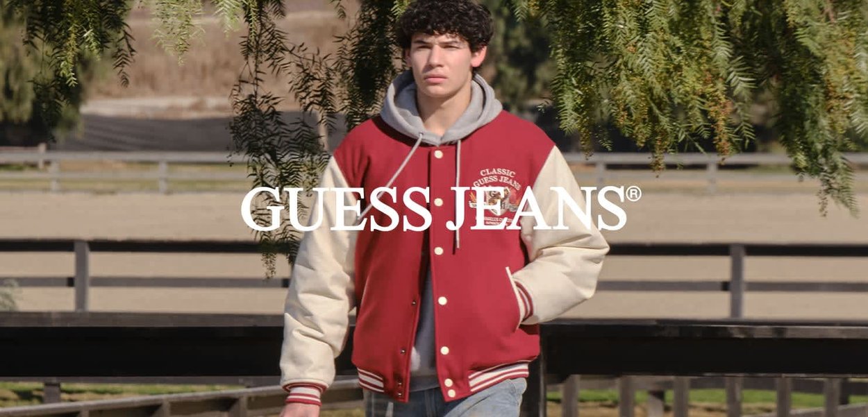 Junger Mann in einer roten und cremefarbenen Collegejacke sowie einem grauen Kapuzenpullover, der im Freien mit Bäumen und einem Holzzaun im Hintergrund geht.