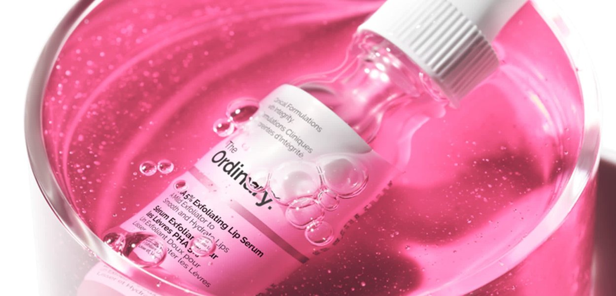 Clear glass dropper bottle with white cap containing pink lip serum, submerged in pink liquid with bubbles, labeled "The Ordinary."