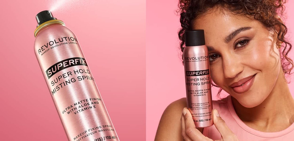 Donna con capelli ricci tiene in mano una bomboletta rosa di Revolution Superfix spray fissante per trucco su sfondo rosa.