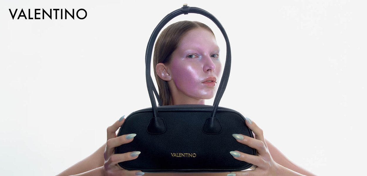 Frau hält eine schwarze Valentino-Handtasche vor ihr Gesicht, das durch die Griffe der Tasche eingerahmt zu sein scheint, mit einem glänzenden rosafarbenen Gesichtshighlight.