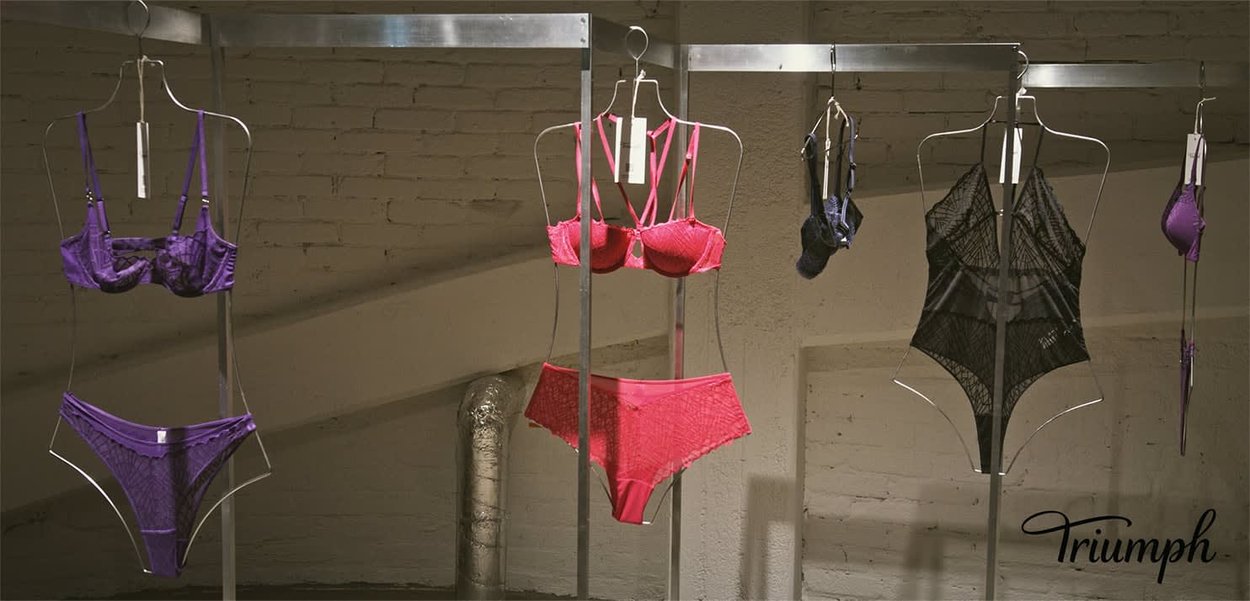 Lila und rosa BHs mit Spitzen- und Netzdetails, ein schwarzes Spitzenbody, auf metallenen Kleiderbügeln vor einem Ziegelhintergrund präsentiert.