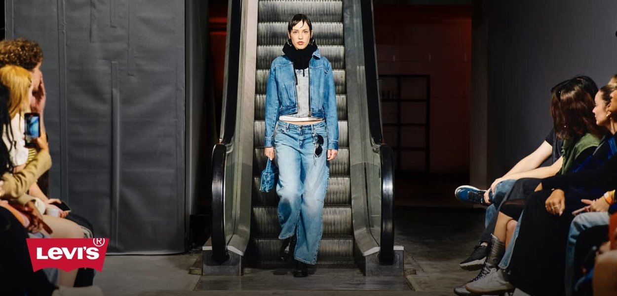 Denim outfit featuring a cropped jacket and wide-leg jeans, both in light blue. Accessories include a matching blue handbag. Escalator background.